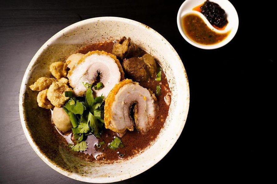 A bowl of dark brown broth is full of crispy chashu pork, meatballs, and herbs.