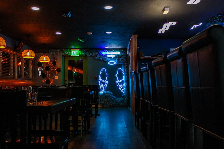 A dimly lit restaurant interior features neon angel wings and a halo under neon words reading "Tequila is cheaper than therapy."