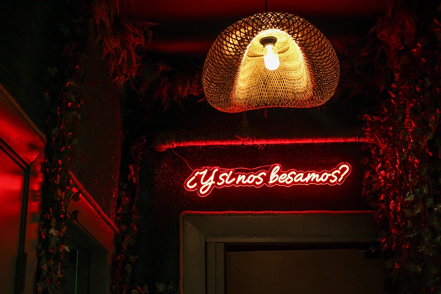 A dimly lit restaurant interior has walls covered with fake leaves and neon signage reading "¿Y si nos besamos?"