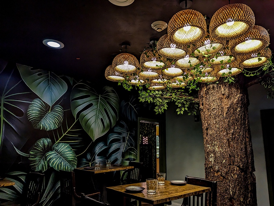 A restaurant dining room features jungle wallpaper and a tree trunk with distinctive basket-like lights coming off of the top with fake leaves.