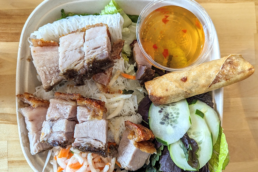 Overhead view of a vermicelli bowl with cucumbers, mint, pickles, fish sauce, egg roll, and thick chunks of pork belly with crispy skin.