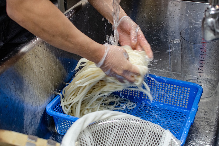 Two gloved hands rinse just-boiled udon noodles in a blue tub in a commercial sink.