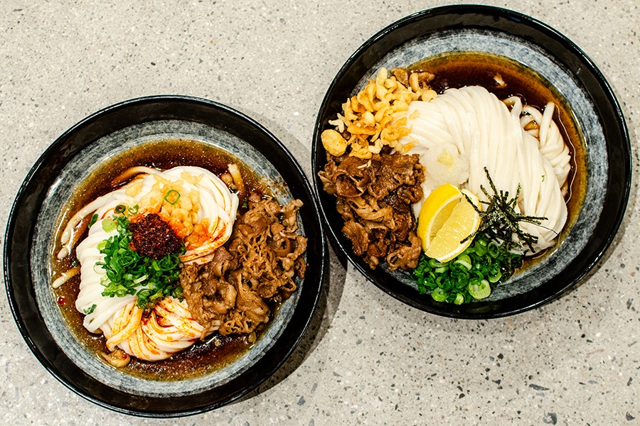 Overhead view of two bowls of udon, each featuring thick noodles, thinly sliced beef, scallions, crispy tempura flakes, and other toppings.