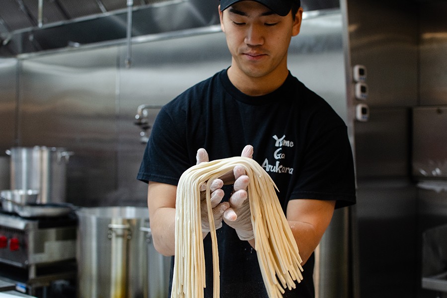 A man in a black t-shirt that says Yume Ga Arukara holds a batch of fresh udon in gloved hands in a restaurant kitchen.