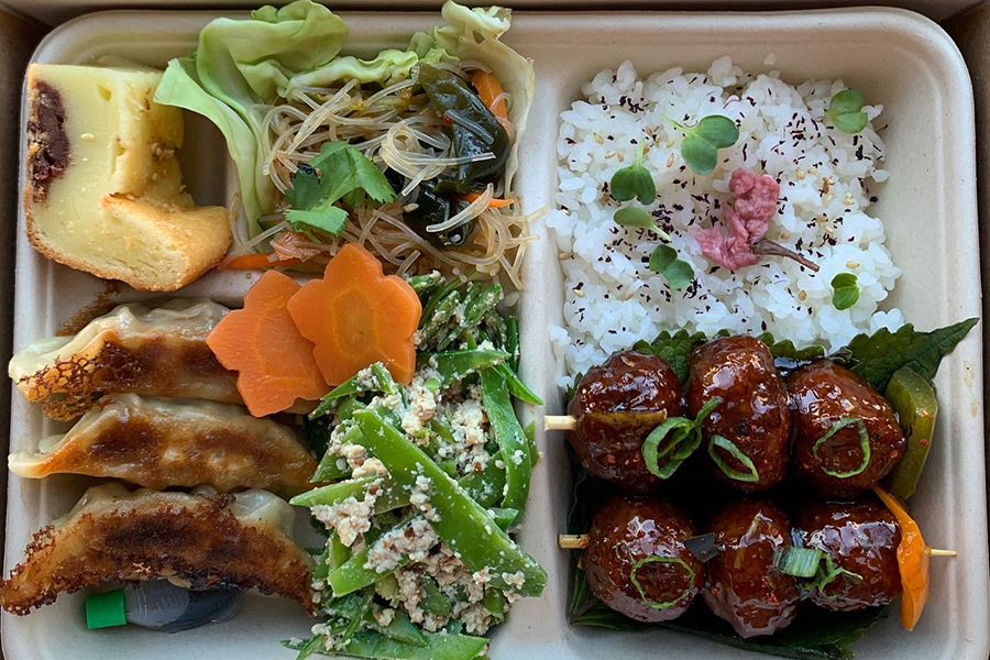 Overhead view of a bento box with seasoned white rice, saucy meatball skewers, gyoza, noodles, butter cake, and more.