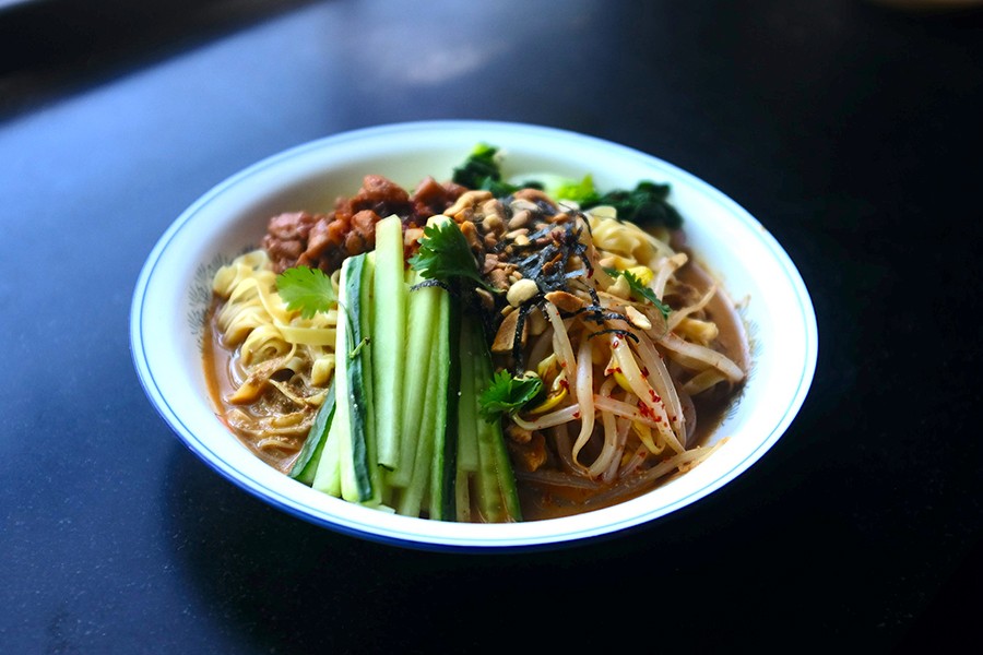 A white bowl is full of noodles, celery, seaweed, peanuts, and more in a brown-orange broth.