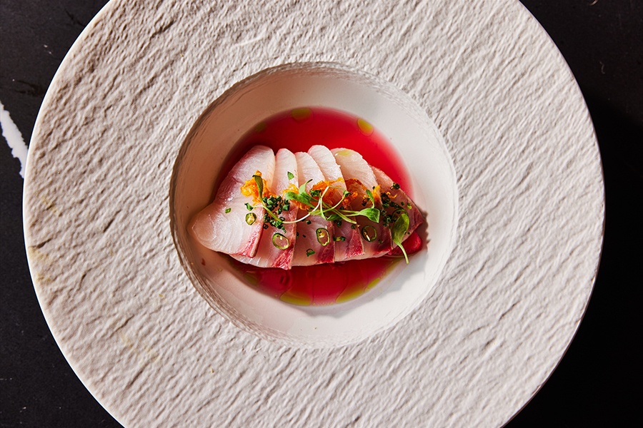 Overhead view of a white plate of sashimi, garnished with greens and sitting in a pinkish thin sauce.