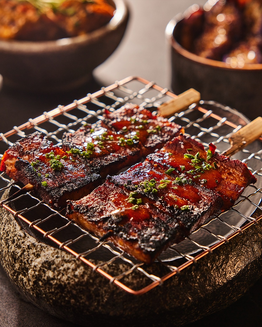 Charred rectangles of pork belly on small wooden skewers sit atop a grate on a small charcoal grill.