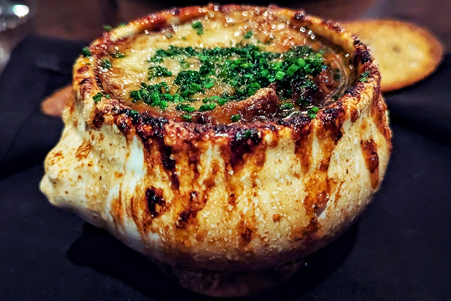 A bowl of French onion soup with charred cheese dripping over the sides of the bowl and thinly sliced chives on top.