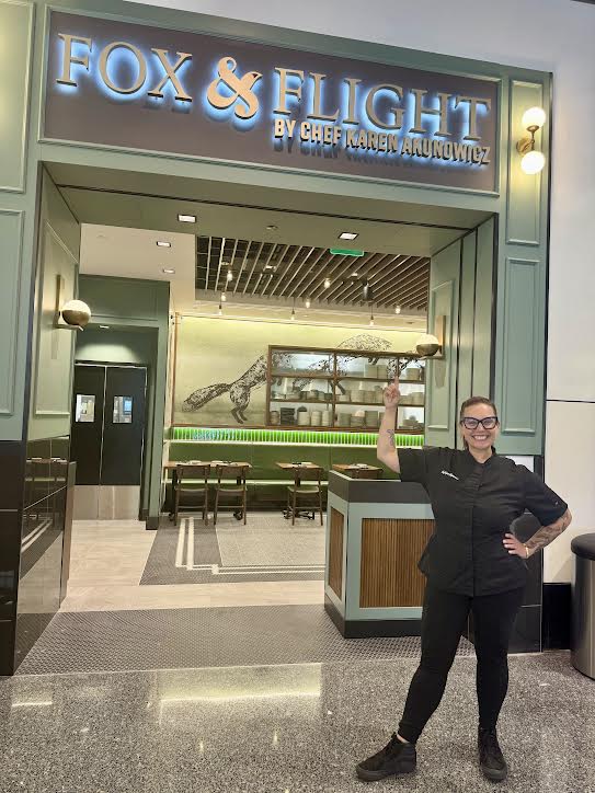 A woman in black clothes and glasses smiles and stands in front of a restaurant, pointing up at the signage, with reads Fox and Flight.