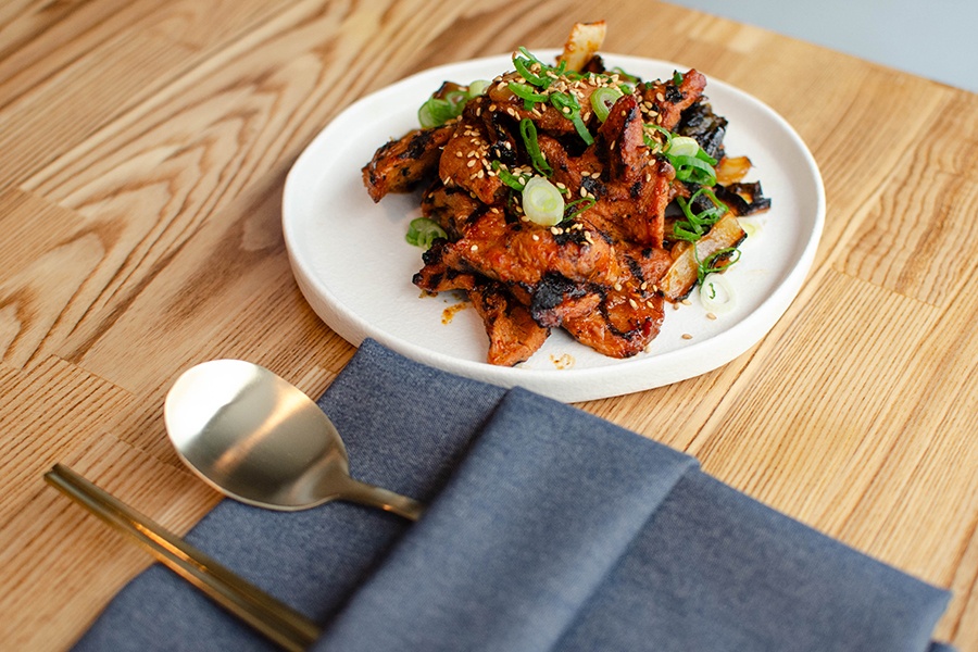 A plate of thin slices of grilled pork with scallions and sesame seeds sits on a light wooden table.