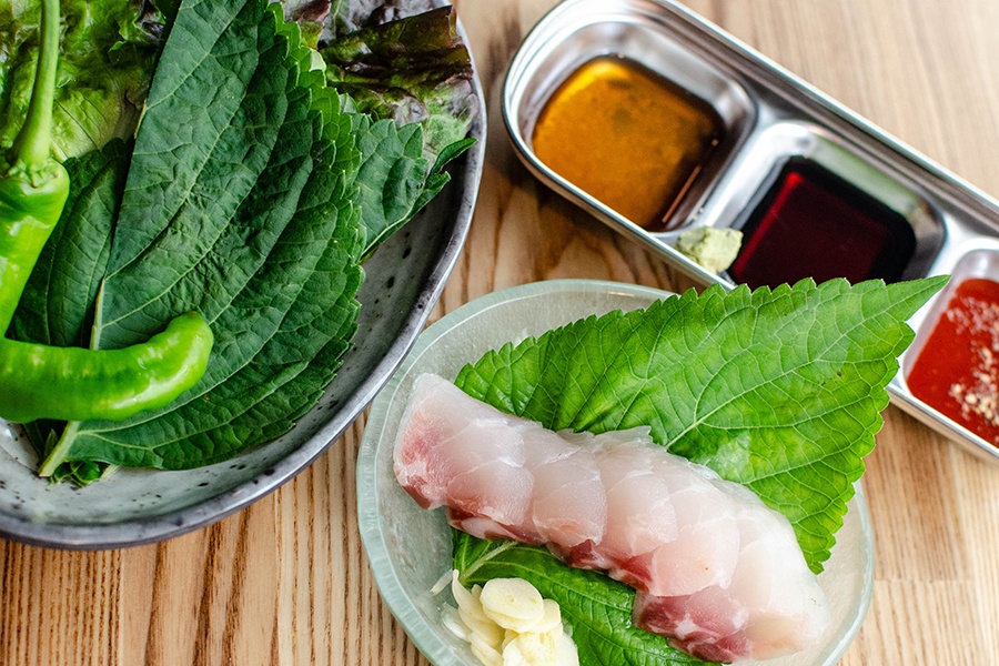 Slices of raw fish are accompanied by three sauces, large leaves, and other garnishes.