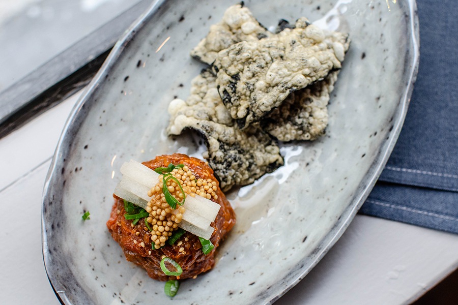 A scoop of orange beef tartare is topped with whole mustard seeds, small sticks of pear, and lightly battered and fried pieces of seaweed.