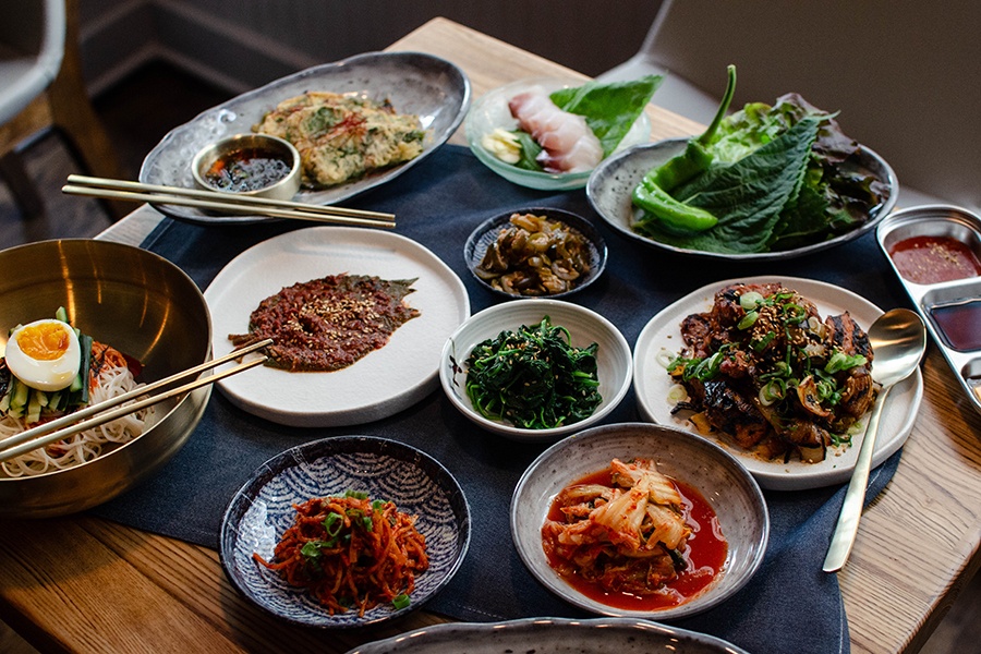 A table is covered with dishes of Korean food, including a selection of small side dishes of kimchi and more.