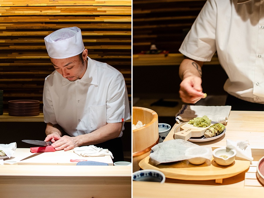 A sushi chef dressed in a white shirt and hat slices raw fish and grabs a bit of fresh wasabi.