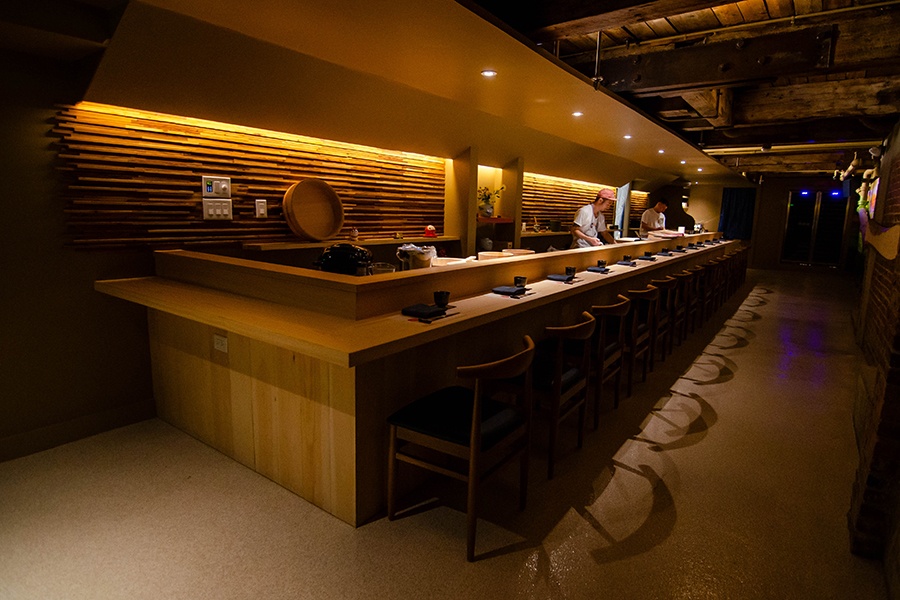 Two restaurant employees work behind a sushi bar in a small, dark basement space.