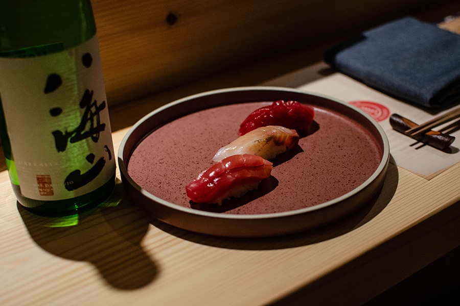 Three pieces of simple nigiri are lined up on a plate with a bottle of sake in the background.