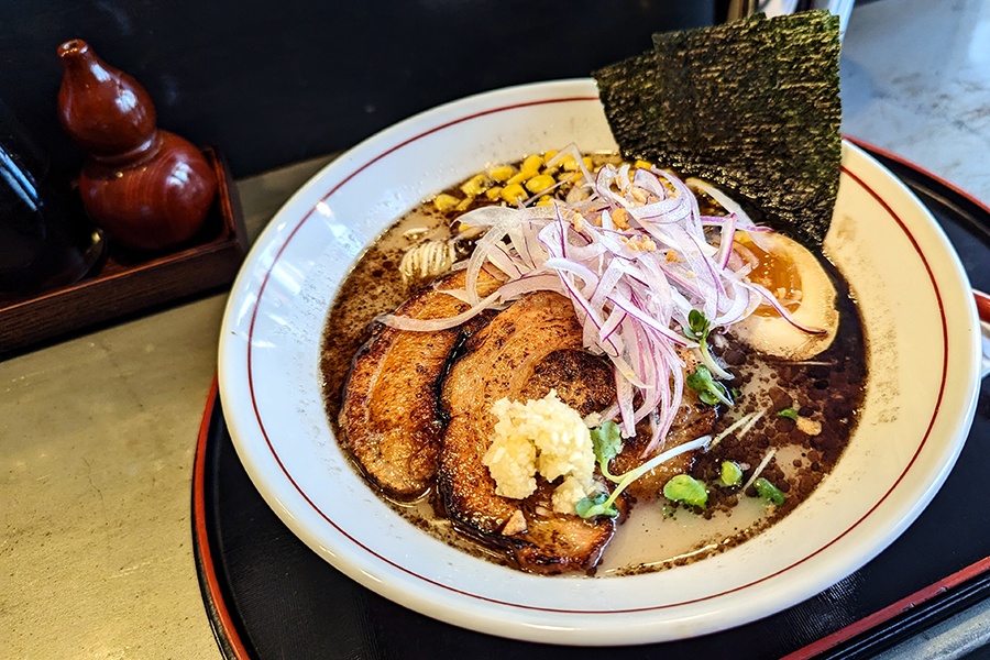 A bowl of ramen features charred pieces of pork, red onion, egg, and corn.