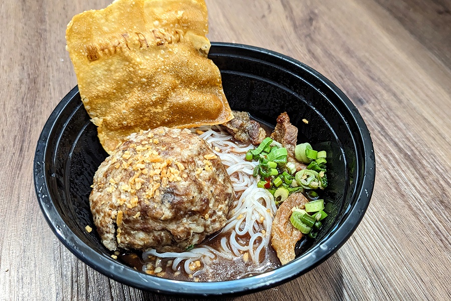 A black plastic takeout bowl is full of broth, thin rice noodles, a crispy wonton strip, and a giant pork meatball.