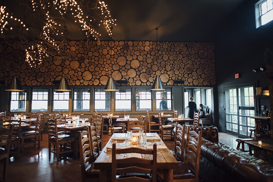 A woodsy restaurant dining room features a wall made of cut logs, brown leather lounge seating, and wooden tables and chairs.