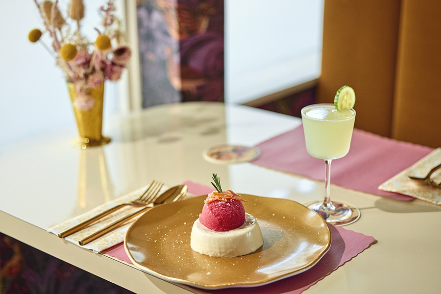 A scoop of pink sorbet sits atop panna cotta with a cucumber-garnished cocktail in the background.