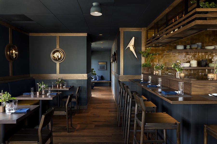 Empty interior of an upscale restaurant with dark blue-gray walls, counter seating by an open kitchen, and light wooden accents.