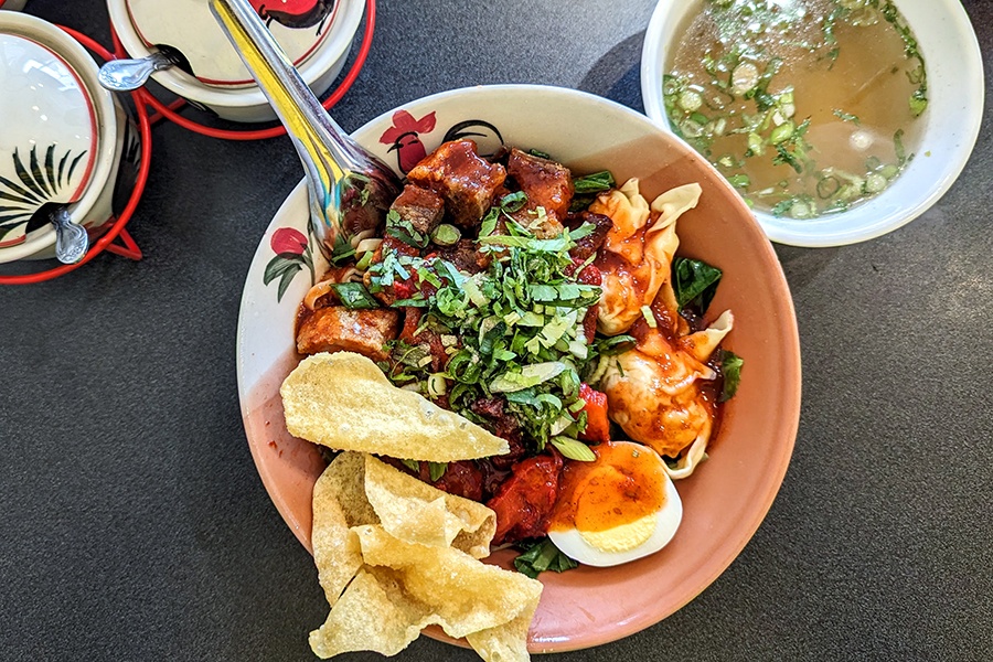 Overhead view of a Thai noodle bowl with pork, eggs, crispy wontons, and greens, with a side of broth.