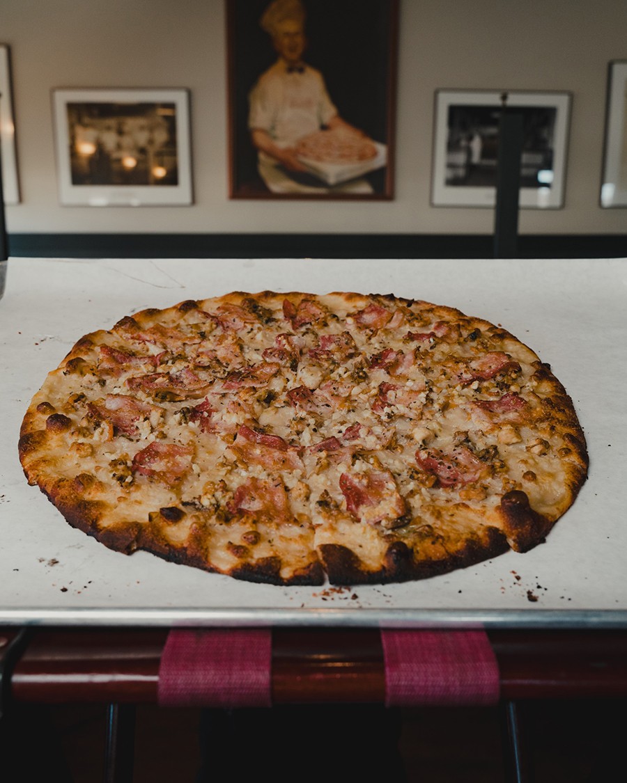 A New Haven-style pizza is visible in front of a painting of Frank Pepe, patriarch of the apizza movement.