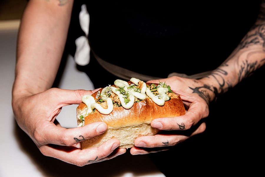 Two hands hold a brioche hot dog-style bun stuffed with fried seafood and topped with a thick squiggle of white sauce.