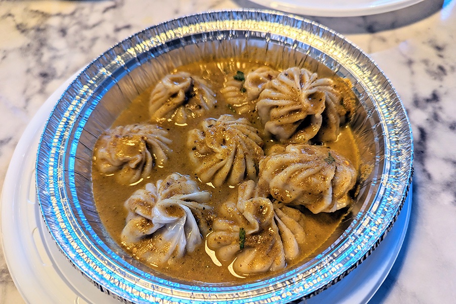 An aluminimum takeout container is full of seven steamed Nepalese dumplings in a thin brownish-orange broth.