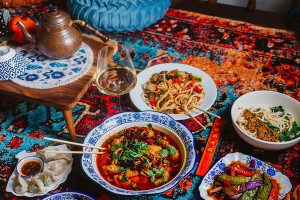 Several Uyghur noodle dishes and a dish with hot chili oil are spread on a colorful carpet with a copper pot of tea and a glass of wine in view.