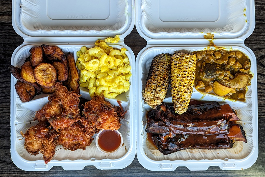 Overhead view of two white plastic takeout containers full of Caribbean food, including fried, breaded shrimp, saucy ribs, mac and cheese, charred corn on the cob, and more.