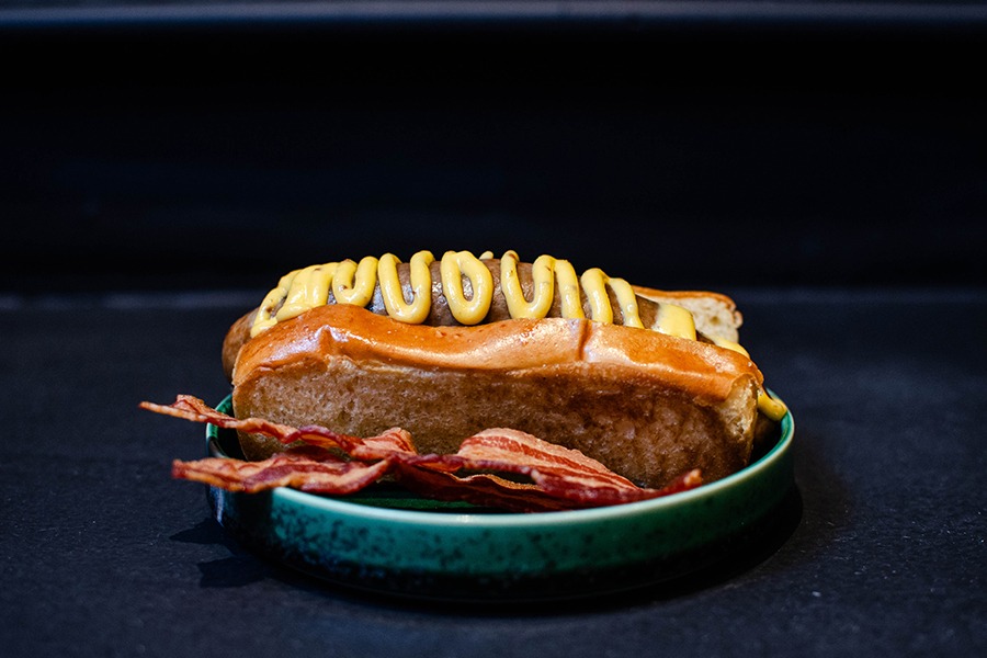 A hot dog is topped with a squiggle of a mustard-like sauce and has a side of crispy bacon strips.