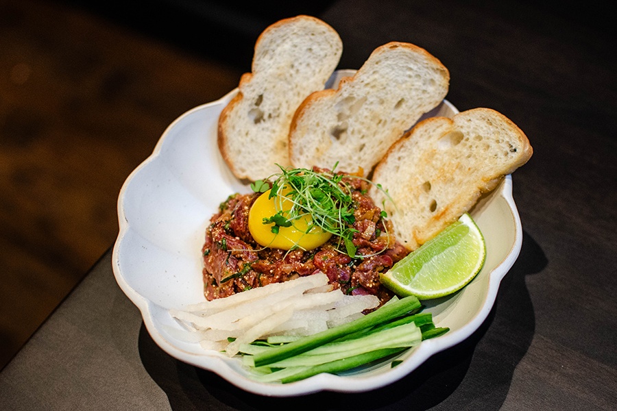 Beef tartare is topped with a cured egg yolk and has sides of thinly slices cucumbers and Asian pears, a lime wedge, and toasted bread.