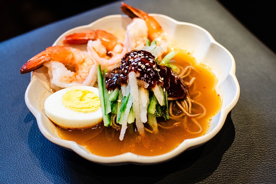 Thin noodles sit in a thin orange broth with a hard-boiled egg, shrimp, cucumber slices, and a thick red chili jam.