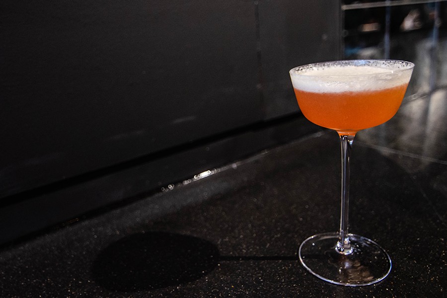 A pink cocktail, topped with foam, sits in a delicate glass on a shiny black bar.