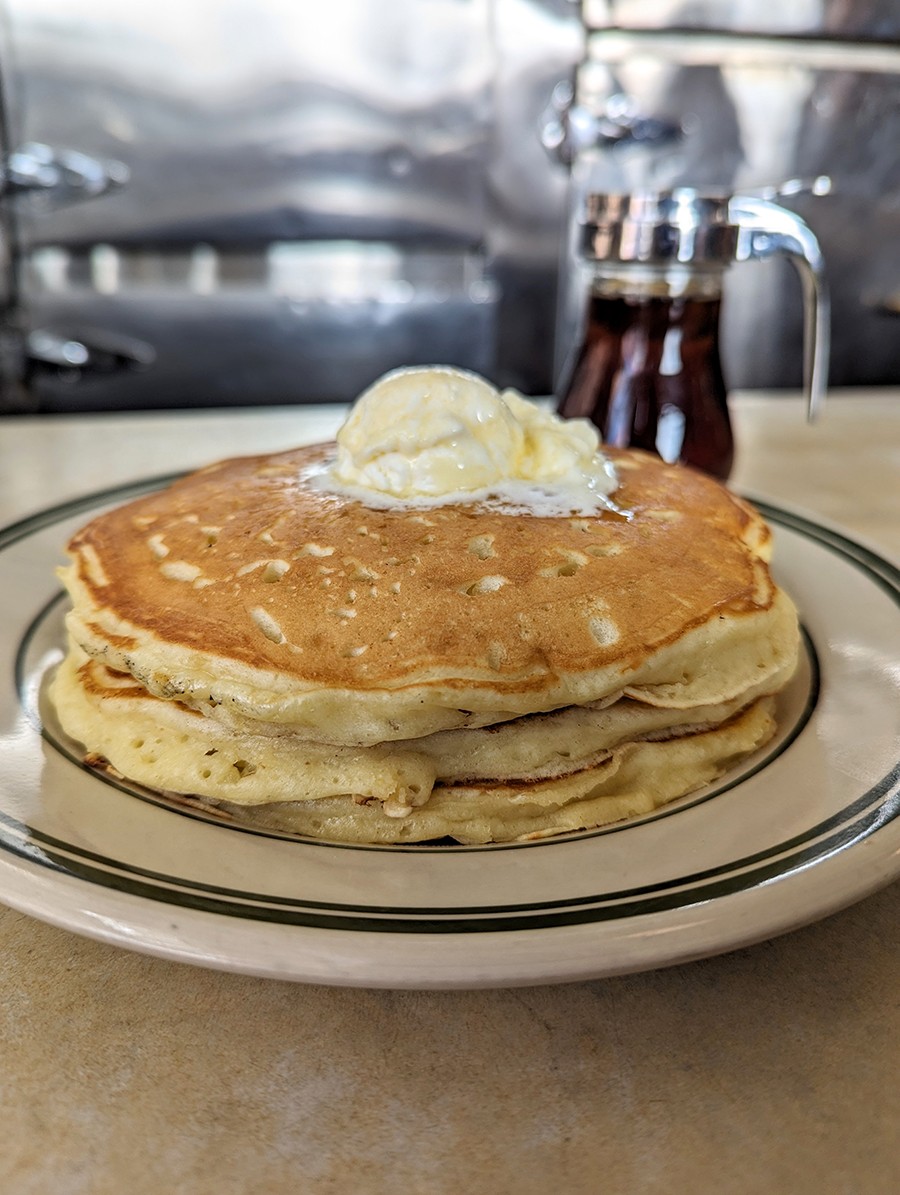 A stack of butter-topped pancakes with a glass container of maple syrup in the background.
