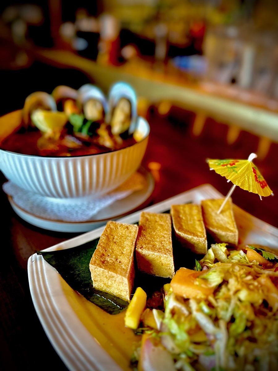 Fried tofu, one piece garnished with a cocktail umbrella, sits in front of a mussel dish on a restaurant table.