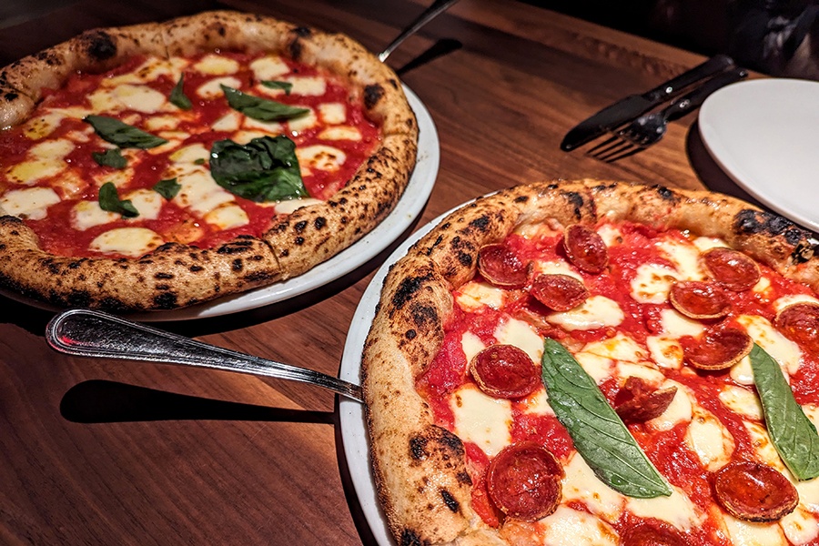 Two Neapolitan-style pizzas, one with cheese, tomato, and basil, and the other with pepperoni, sit on a wooden table.