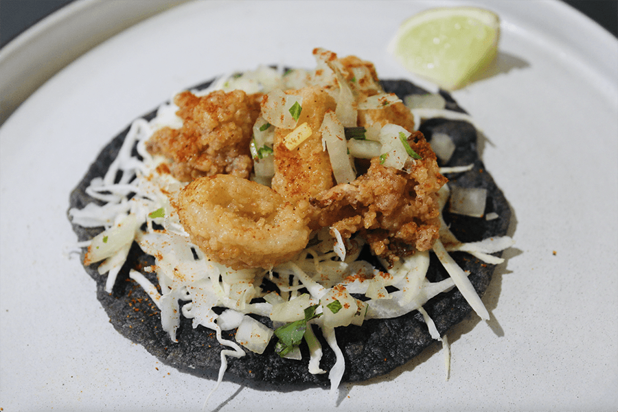 Fried, breaded calamari sits atop a bed of shredded cabbage on a black tortilla, with a wedge of lime accompanying.