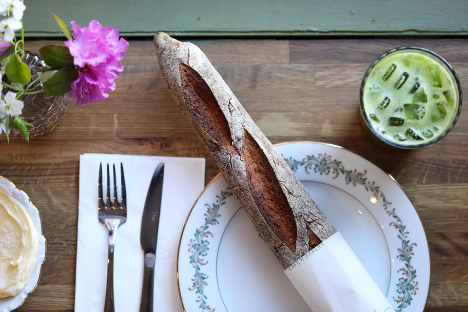 A long, skinny baguette on a plate with an iced green drink, silverware, and a pink flower.