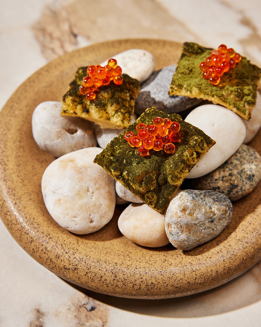 Little fried dough squares covered in dark green powder and orange roe are displayed on stones on a round plate.