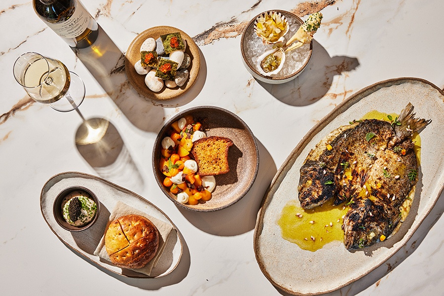 Overhead view of upscale Greek dishes on a marble background, including a whole grilled fish, raw shellfish, a focaccia-like bread with a caviar-topped spread, and more.