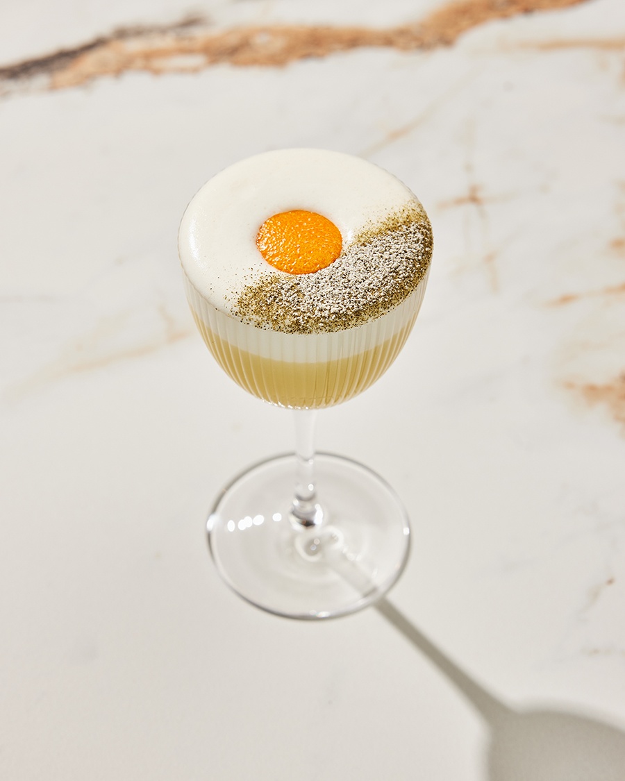 A yellow cocktail is topped with foam, spices, and a circle of mandarin orange skin.