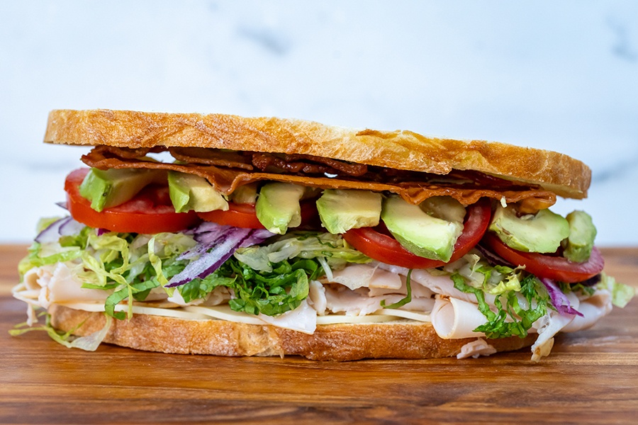 Turkey, lettuce, red onion, tomato, avocado, and bacon are stacked between two long slices of bread.