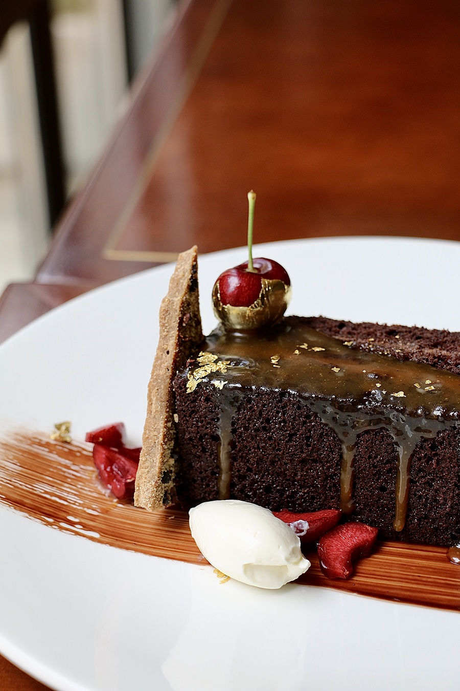 A slice of chocolate cake drips with caramel and is garnished with gold flakes and a cherry.