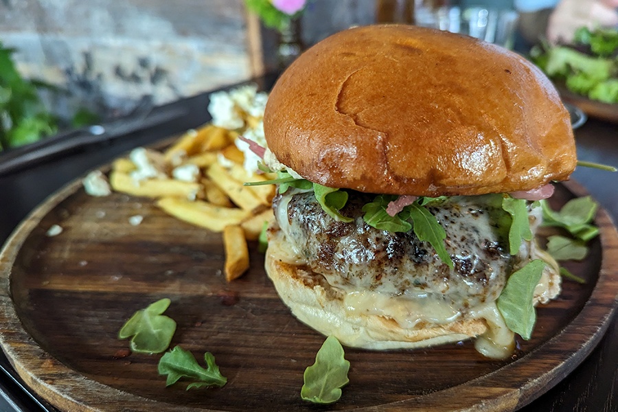 A burger is topped with melted cheese, arugula, and pickled shallots and is displayed on a wooden plate.