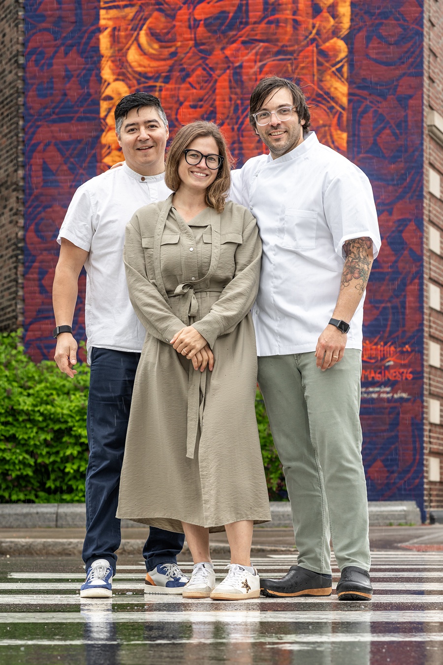 Two men in chef's whites and a woman stand smiling in a crosswalk in front of a colorful mural in Central Square, Cambridge, Massachusetts.