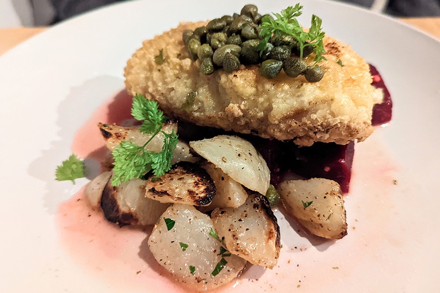 A breaded piece of fish is topped with capers and has a side of roasted potatoes.