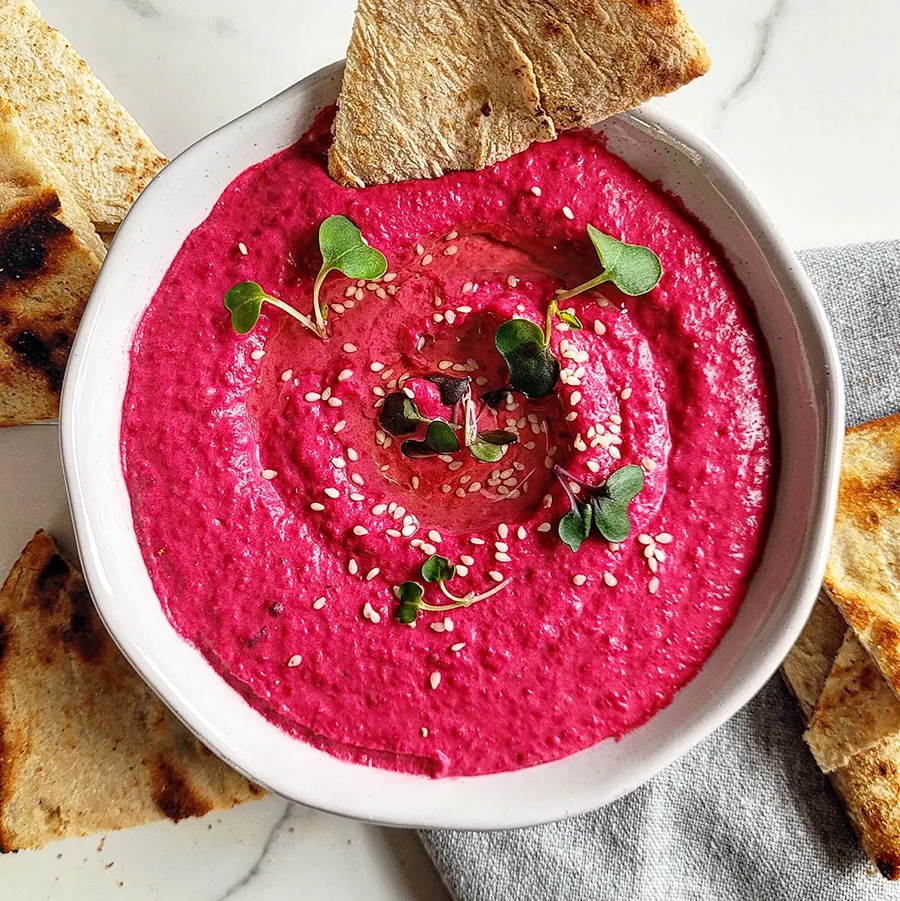 Bright pink hummus topped with sesame seeds and microgreens, accompanied by crispy pita.